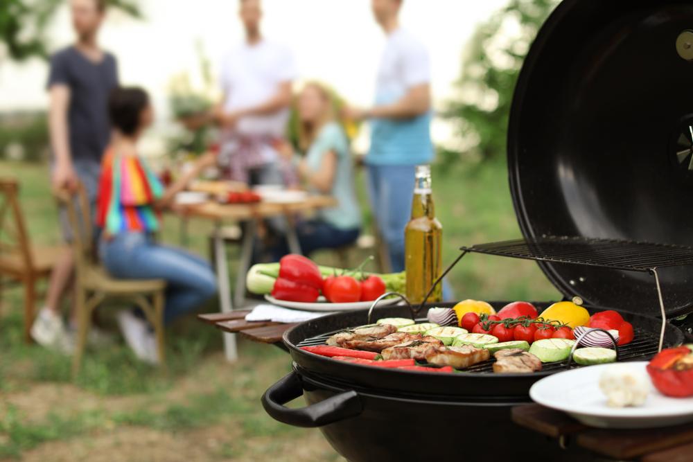 A grill with meat and vegetables cooking, with a small gathering of people at a picnic table in the background.